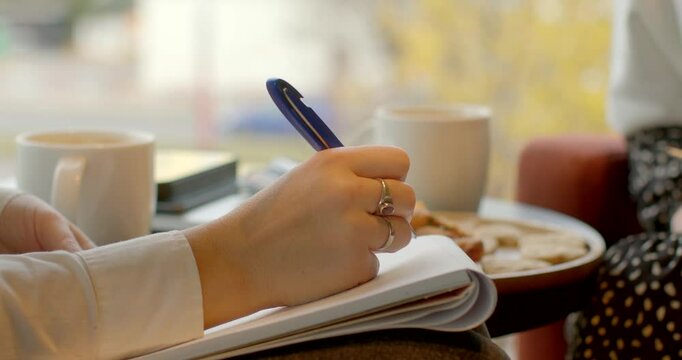 An individual is taking notes while enjoying coffee and breakfast items on the table nearby