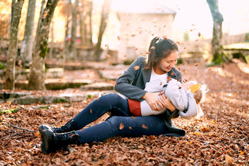Smiling mom hugging little girl on her knees while sitting in autumn park under falling leaves