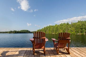 The calm blue lake in Muskoka reflects the summer sky, surrounded by lush trees. Two Adirondack...