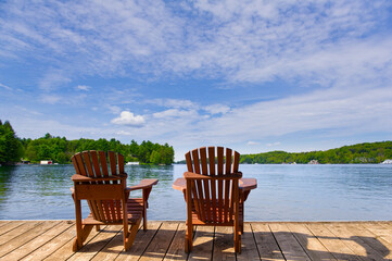 The calm blue lake in Muskoka reflects the summer sky, surrounded by cozy cabins nestled among the trees. Two Adirondack chairs sit on a wooden dock, facing the water.