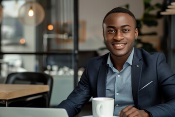 Fototapeta premium Happy smiling male sitting at desk in coworking office, drinking coffee from takeaway cup and doing work on his laptop, looking at camera