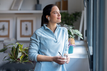 Beautiful sporty woman eating yogurt while listening music with earphone standing near the window...