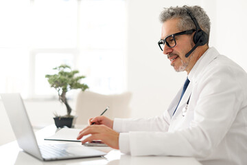 A grey-haired doctor is confidently using a laptop for a medical consultation, his headset and...