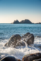 Sea waves hitting and splashing against a rocky shore forming different shapes with an island and a clear blue sky in the background