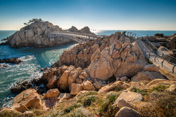Rocky cliffs of the sea of Japan coast in Korea with a pedestrian bridge leading to an island with clear blue sky and the sun 