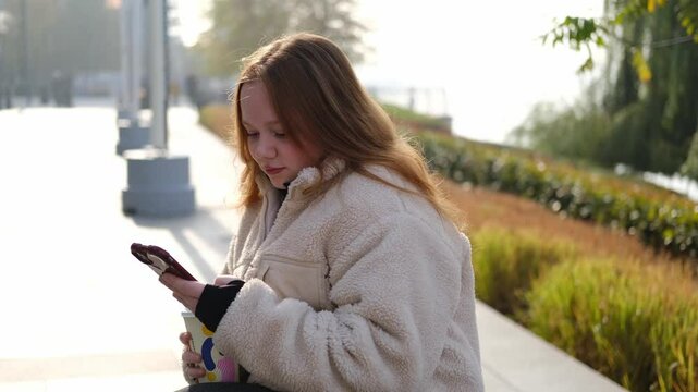 A young red-haired woman in a beige fleece jacket is drinking coffee sitting on a bench by the river and chatting on the phone, against the background of urban landscape