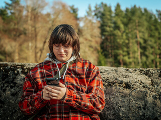 A man in a red plaid jacket and glasses looks at his smartphone while leaning against a stone structure. The natural backdrop of trees and warm sunlight add to the relaxing outdoor setting.