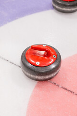 Close-up of a red coloured curling stone sliding on ice of stadium, placed near concentric rings and other stone in the background