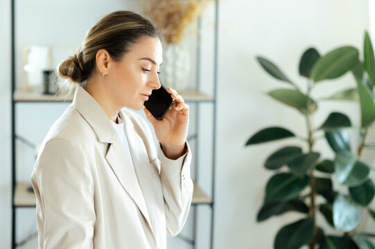 Focused caucasian businesswoman in a beige blazer stands in the office while talking on her smartphone, handling important business matters with a serious facial expression