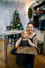 Little girl stands near her mother squatting with a tray of raw Christmas cookies