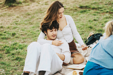 A young couple sits closely on a picnic blanket, enjoying a sunny day outdoors. They are surrounded...