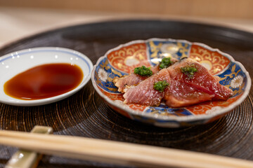 Seared bonito sashimi garnished with fresh green sauce, served on an ornate ceramic plate with soy sauce on the side, showcasing the elegance and minimalism of Japanese cuisine