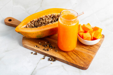 A halved papaya, vibrant smoothie in a glass jar with a straw, fresh cubes in a bowl, and scattered...