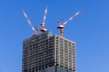 Three towering construction cranes operate atop a partially built skyscraper covered with...