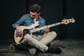 A musician plays a bass guitar while seated on the floor of a studio. The setting emphasizes...