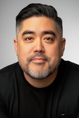 Obraz premium A man with a neatly groomed beard and modern hairstyle looks directly at the viewer, displaying a composed expression. His black shirt adds to the simplicity of the neutral background