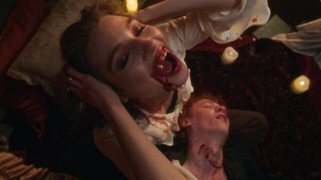 High angle portrait shot of sinister vampire woman with blood-stained chin and fangs holding lifeless head of man with bite marks on his neck in dimly lit, candlelit bedroom