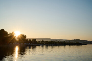 Naklejka premium Gentle sunrise casts golden light on tranquil water, illuminating the shoreline and distant hills.