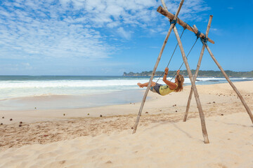 Happy girl have fun swinging high in mid air. Flying on rope swing on sea beach. Travel adventure on paradise tropical island. Family lifestyle, activity on summer vacation.