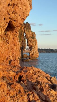 High rocky cliffs with a natural sea arch formed in orange limestone. Calm blue sea water and distant city coastline under a clear sky. Golden hour sunlight on porous rock texture.
