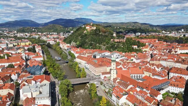 Aerial view of historic old town with uhrturm clocktower and red roofs surrounded by mountains and greenery, Graz, Styria, Austria.