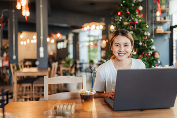 Asian woman in casual white shirt, working remotely on laptop in cafe, The background festive atmosphere with decorative hanging lights and Christmas tree. Blend of work and lifestyle.