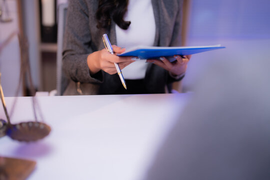 Female lawyer reviewing legal documents and holding a pen, sitting at her desk with scales of justice in the foreground, symbolizing law, justice, and legal proceedings