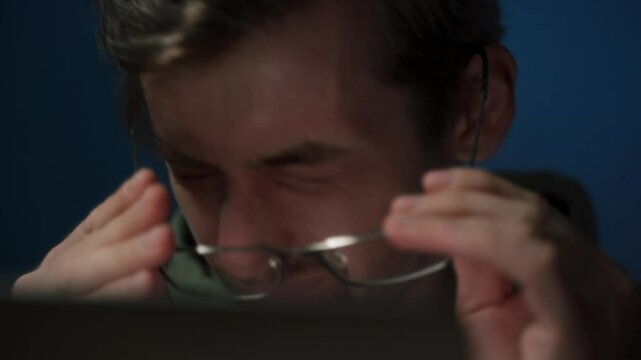 A tired coder wearing eyeglasses working late at night feels a headache and rubs his eyes from fatigue. Overwork concept