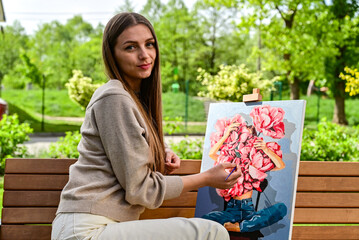A young female artist holding a brush, painting a colorful artwork on canvas in a serene outdoor nature setting.