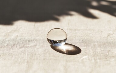 Close-up of a transparent glass sphere casting a bright reflection on a light beige textured surface, illuminated by sunlight creating shadows