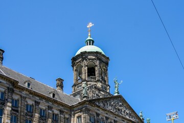 Fototapeta premium A low-angle shot of the Royal Palace of Amsterdam, showcasing its distinctive dome, spire, and facade against a clear blue sky.