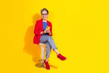 Stylish mature woman in a vibrant outfit holding a smartphone while seated against a bright yellow background