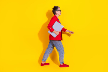 Fototapeta premium Fashionable senior woman walking confidently while holding a laptop against a vibrant yellow background showcasing elegance and style