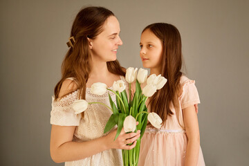 A mother and her daughter stand closely together, exchanging smiles while holding a bouquet of white tulips. Their outfits are soft and light, adding to the warm atmosphere