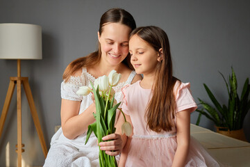 Obraz premium A mother and her daughter sit together on a sofa, smiling as they admire a bouquet of white tulips. Natural light fills the room, creating a warm atmosphere