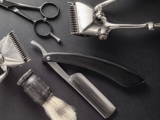 Various vintage barber tools are neatly arranged on a dark surface. The collection includes hair clippers, scissors, a straight razor, and a shaving brush, highlighting classic grooming techniques