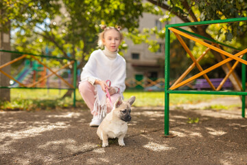 Girl with two ponytails in white and pink clothes squatting next to her small dog
