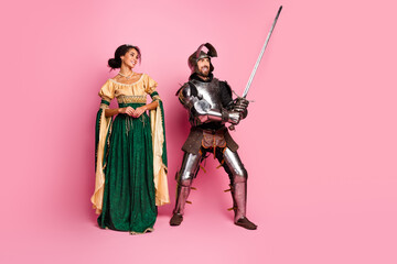 Naklejka premium Playful couple in medieval costumes with knight armor and princess gown against bright pink backdrop