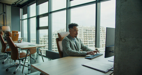 A professional businessman working diligently on a computer in a sleek, modern office with large...