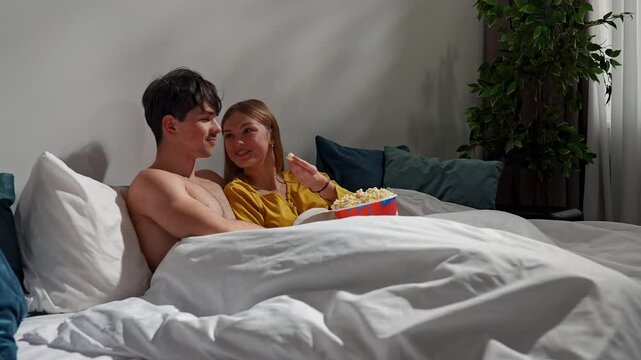 Couple relaxing together in bedroom at home, man and woman laying in bed watching movie and feeding each other with popcorn, kissing and cuddling. Slow motion
