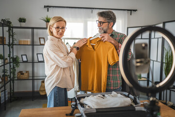 Naklejka premium A mature senior man and woman stand in a cozy, modern living room, proudly showcasing a sweater in front of a camera setup, exuding confidence and joy