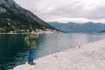 Fototapeta premium Man with a backpack stands on a pier by the sea and photographs the mountains. Back view