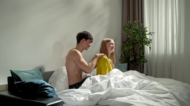 Couple relaxing together in bedroom at home, man and woman sitting in bed, man massaging woman's back and gently touching.