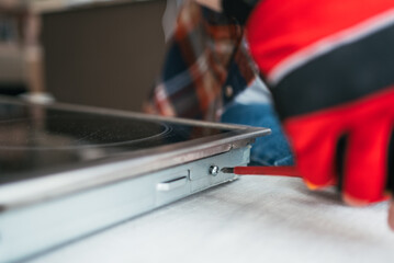 Gloved hand of repairman tightening screw on a ceramic hob during maintenance work in a kitchen,...