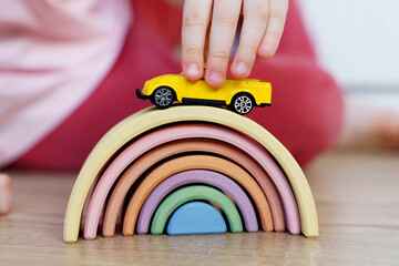 Wooden toy rainbow arc in pastel colors and child's hand holding toy yellow car on top. Natural...