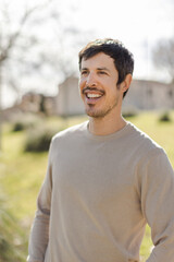 Cheerful middle-aged Caucasian man smiles while gazing into the distance, enjoying the moment. Warm sunlight enhances the peaceful outdoor. Vertical, soft-focus background with trees and building