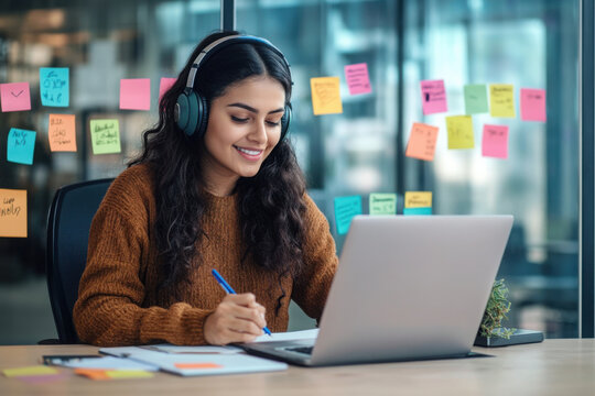 Woman watching online video course, training conference inside office at workplace. Office worker in headphones using laptop for remote meeting, writing data in notebook.