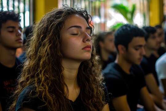 A young woman with curly hair and freckles, eyes closed, seemingly meditating or practicing mindfulness in a group of other people with eyes closed.