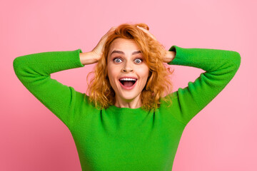 Excited woman wearing green sweater with hands on head in front of bright pink background,...