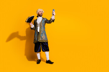 Young man in vintage aristocratic costume using smartphone on yellow background